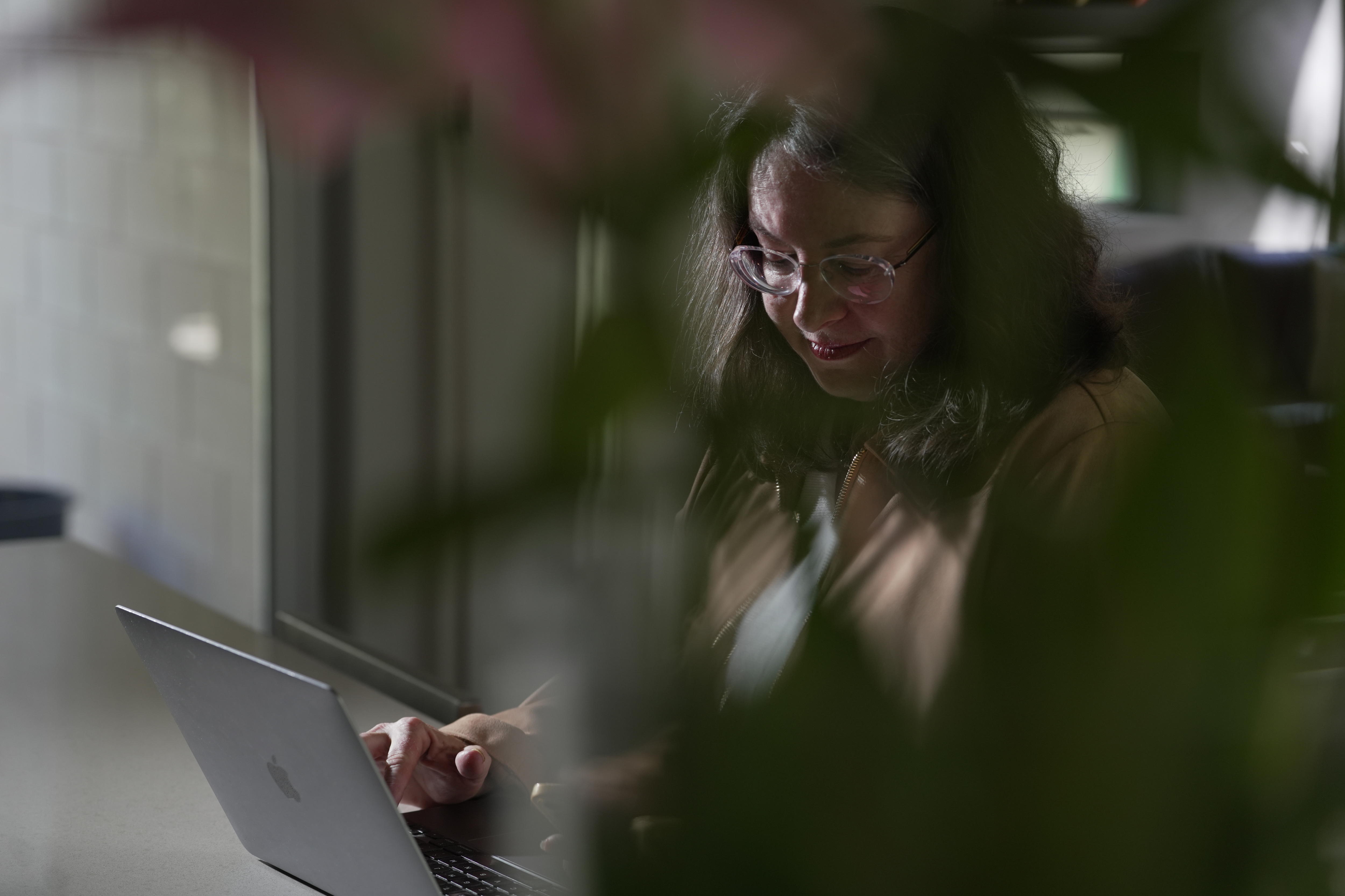 Uma mulher olha para um laptop com uma planta em primeiro plano.