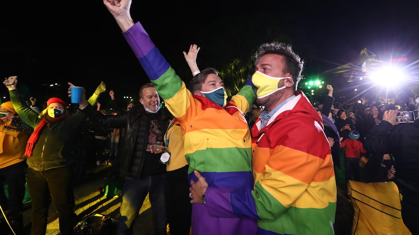 Two people in rainbow coats and green and gold masks cheer.