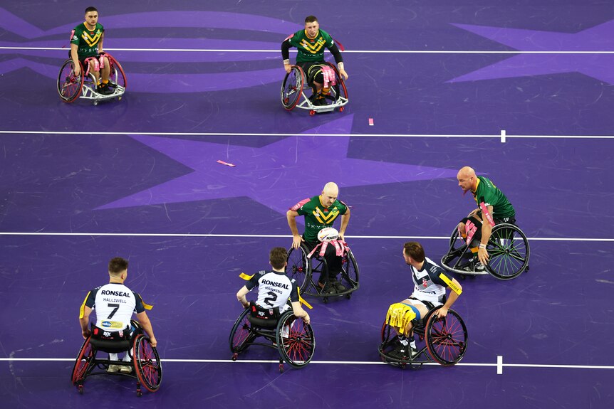 An Australian wheelchair rugby league player holds the ball in his lap as he wheels his chair at the English defensive line.