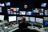 Director wearing headset sitting at desk with numerous panels and monitors in front while news goes to air.