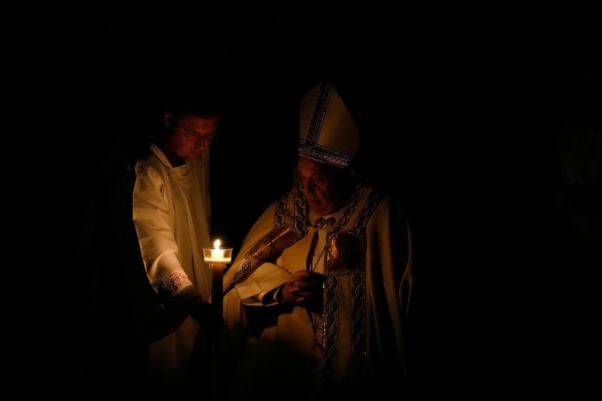 Pope Francis sits in a darkened room, looking at a candle being lit. 