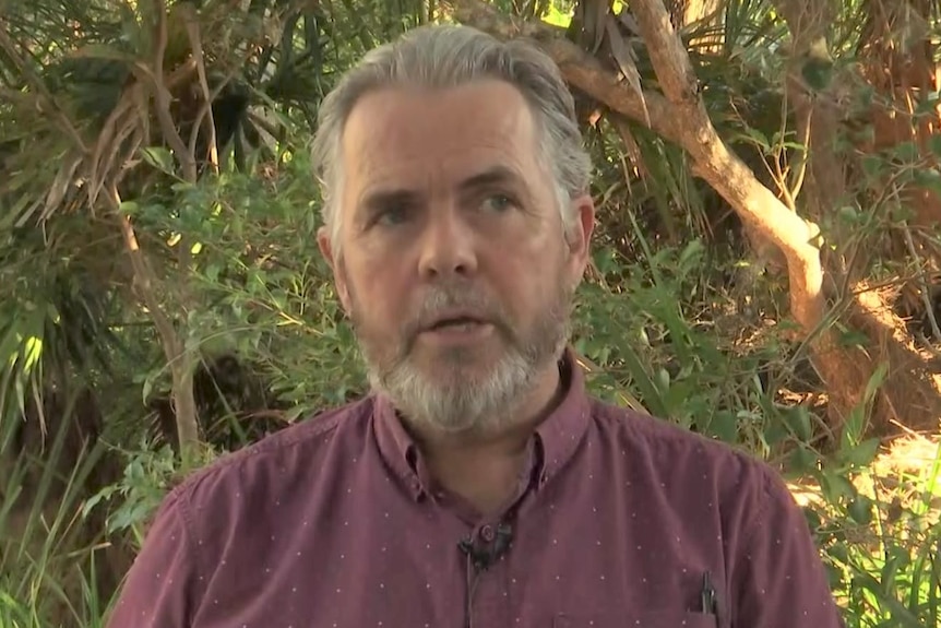 man with short grey hair and beard wearing short sleeved maroon top, hands folded in front of him, standing amongst leafy scrub