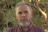 man with short grey hair and beard wearing short sleeved maroon top, hands folded in front of him, standing amongst leafy scrub