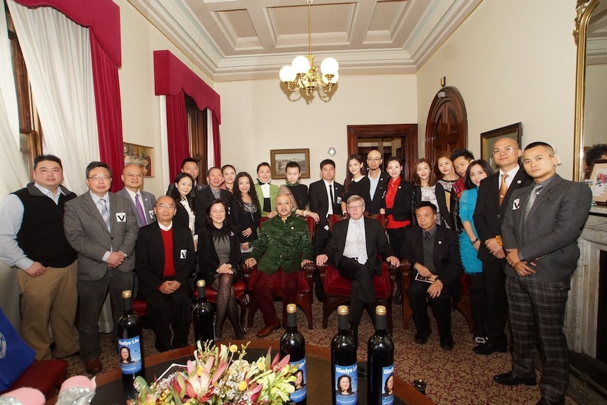 An ornate room with 25 people in semi-formal attire, with four people seated on red velvet chairs.