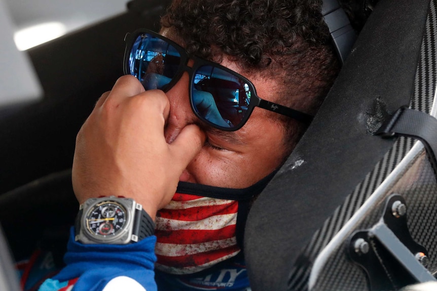 Bubba Wallace holds his hand to his eyes, pushing his blue sunglasses up his forehead, while sitting in a NASCAR seat