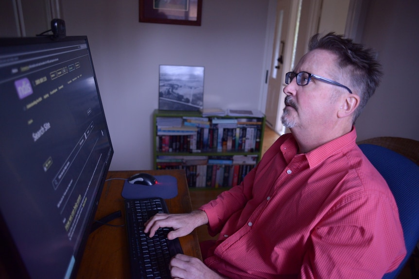 A man wearing glasses sits at a desk and looks at a computer screen.