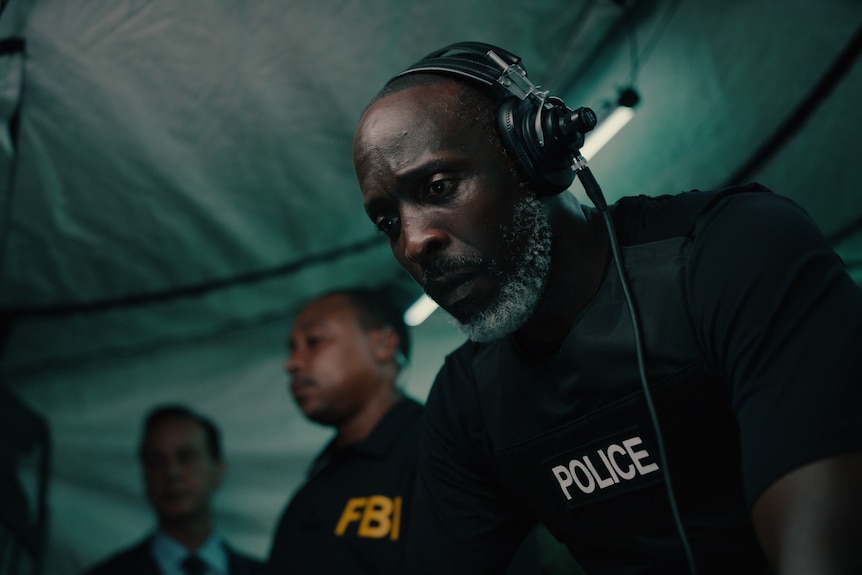 Movie still: Man in police jacket talks into headset.
