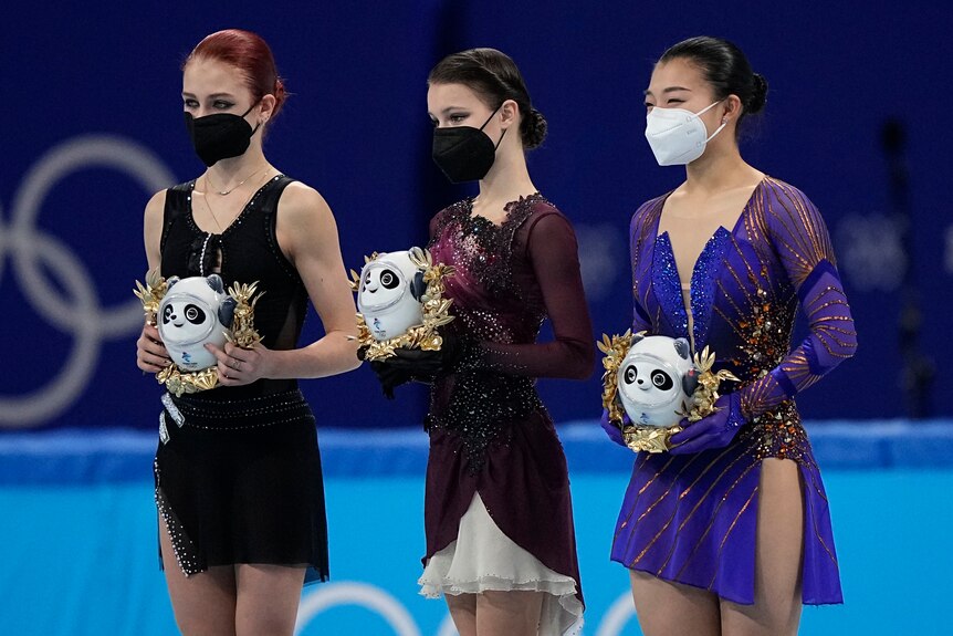 Three female figure skaters - the gold medallist in the middle - stand with masks on holding a Winter Olympic mascot in Beijing.