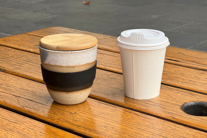 A keep cup with a wooden lid and a white plastic disposable coffee cup sit side-by-side on a wooden table.