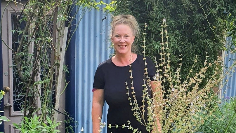 Smiling woman stands at house door surrounded by gardens in a story about finding happiness even in challenging times.