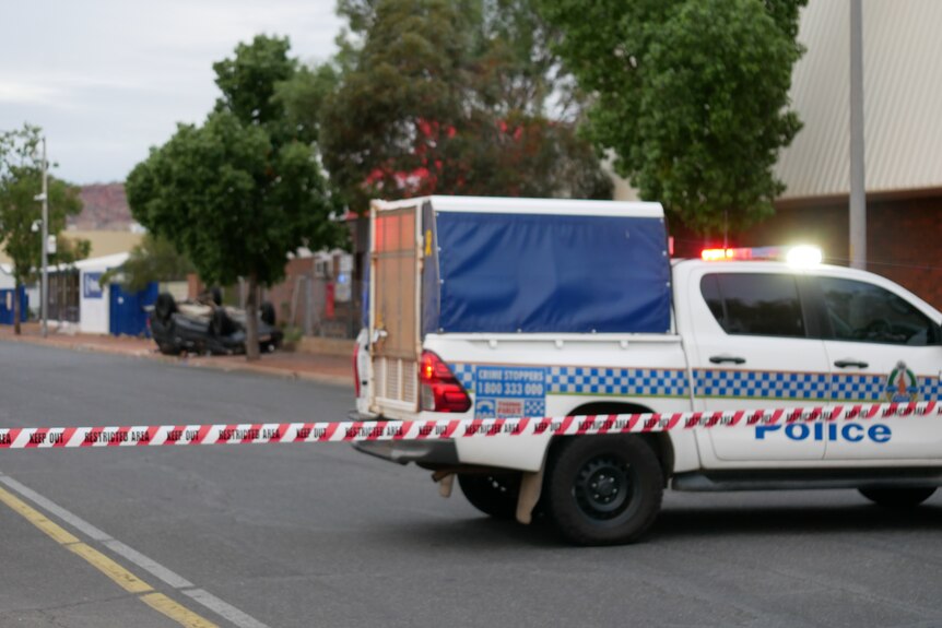 A police wagon parked on a cordoned-off street where az dark-coloured car is lying on its roof.