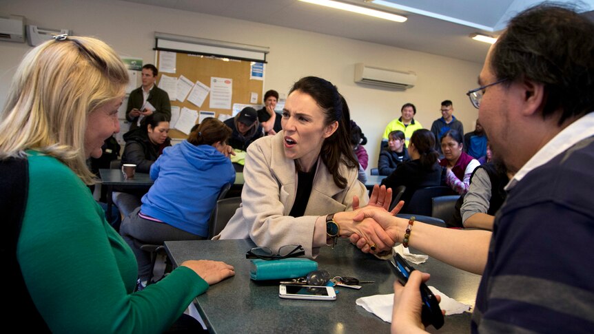 Jacinda Ardern sits at a table with two mushroom factory workers, shaking hands with one and talking to another.