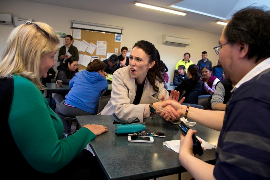 Jacinda Ardern sits at a table with two mushroom factory workers, shaking hands with one and talking to another.