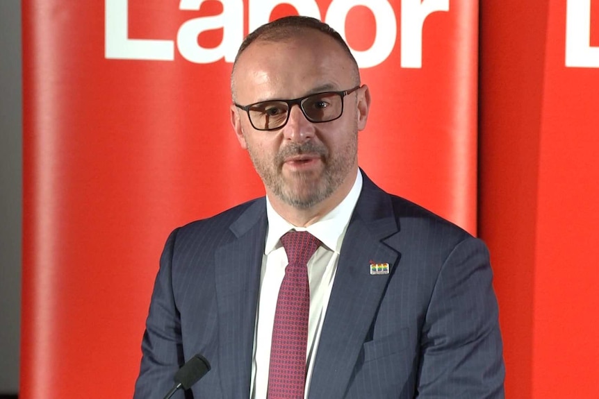 A man at a podium in front of a red banner.