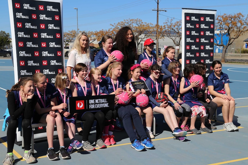 A group of young women in matching blue and pink netball uniforms pose for a team photo, smiling.