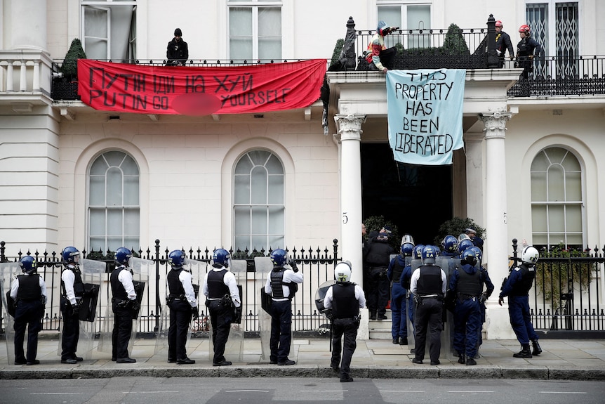 A line of police officers outside a white mansion line up at the door as people hold signs from a balcony above.