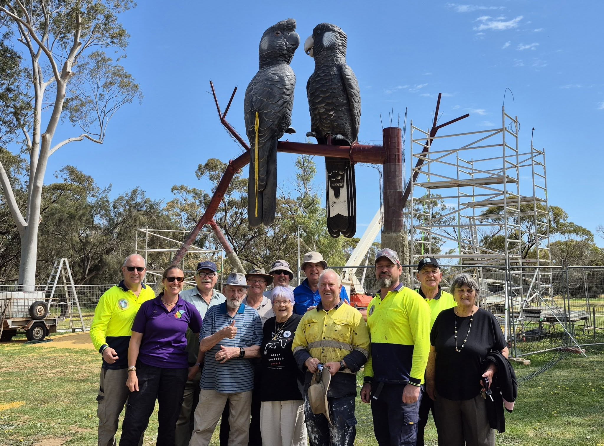A group of people smile in front of a large sculpture of two black cockatoos perched on a branch.
