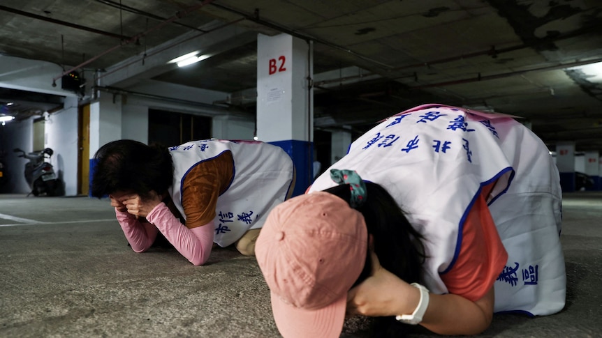 Two people crouch down facing the ground in an undercover car park. 