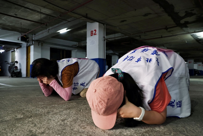 Two people crouch down facing the ground in an undercover car park. 