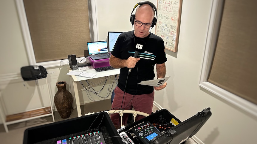 A man with headphones on speaks into a microphone with two large audio panels in front of him