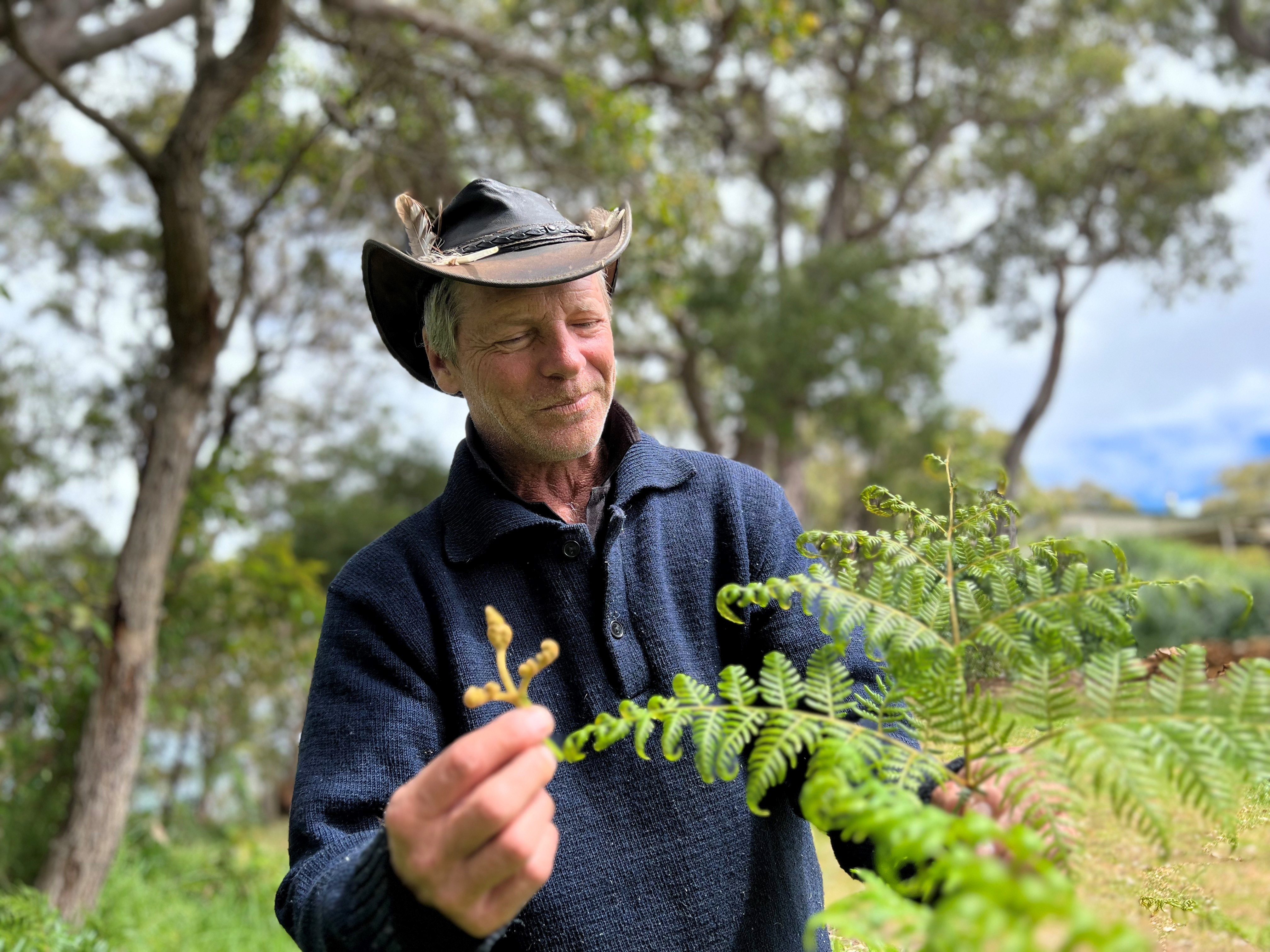man wears a hat and blue jumper holds a green leafy plant and smiles