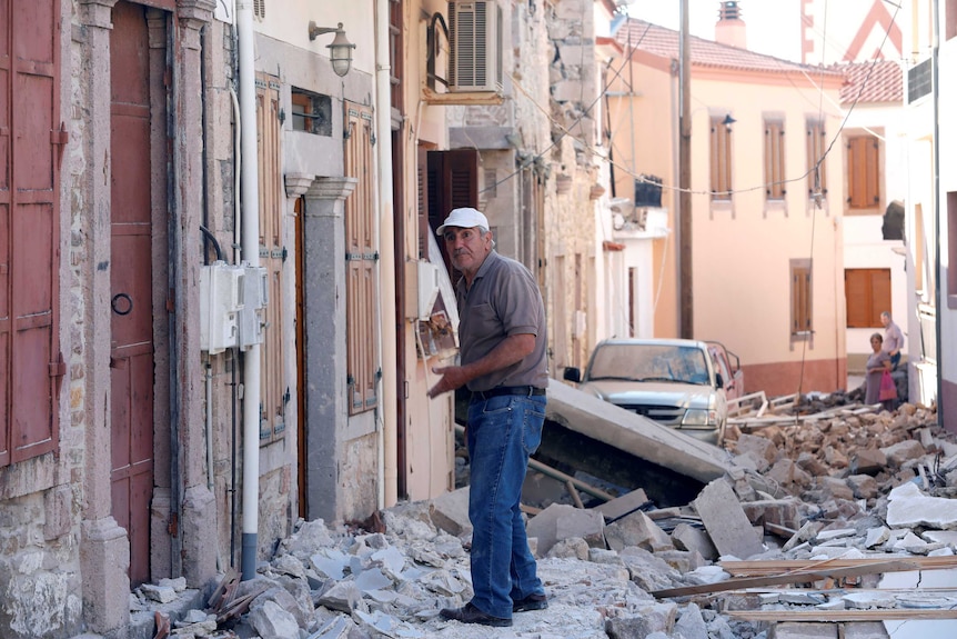 A man stands among damaged buildings, the road covered in rubble