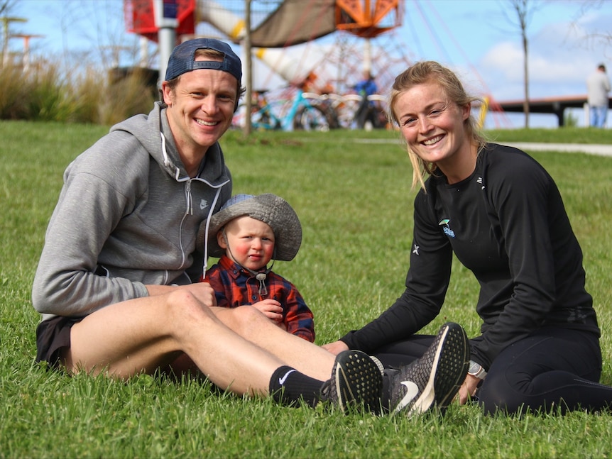 A young blonde couple with their baby sitting on the grass smiling at the camera