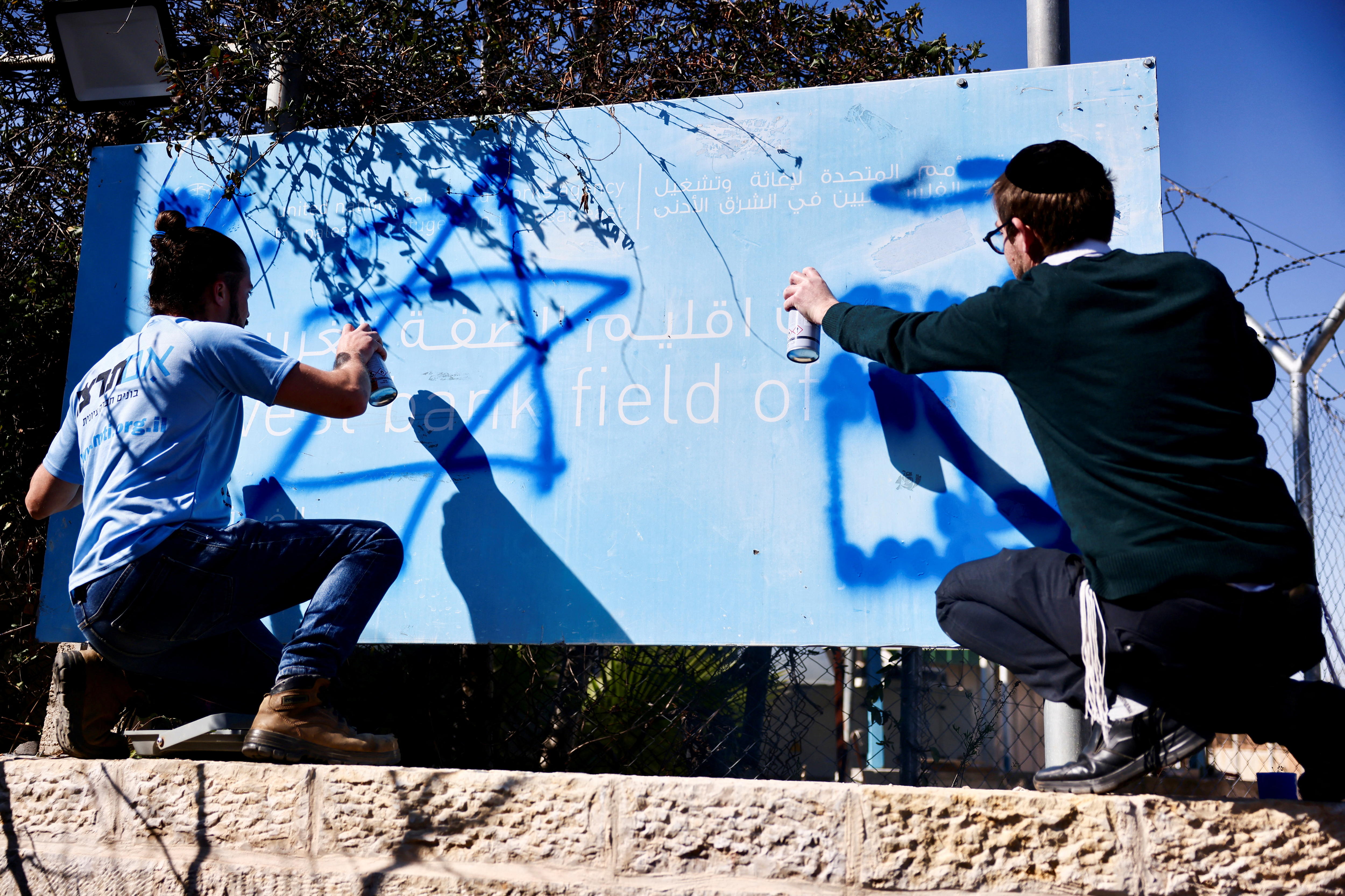 Dois homens pintam com spray uma placa da UNRWA.