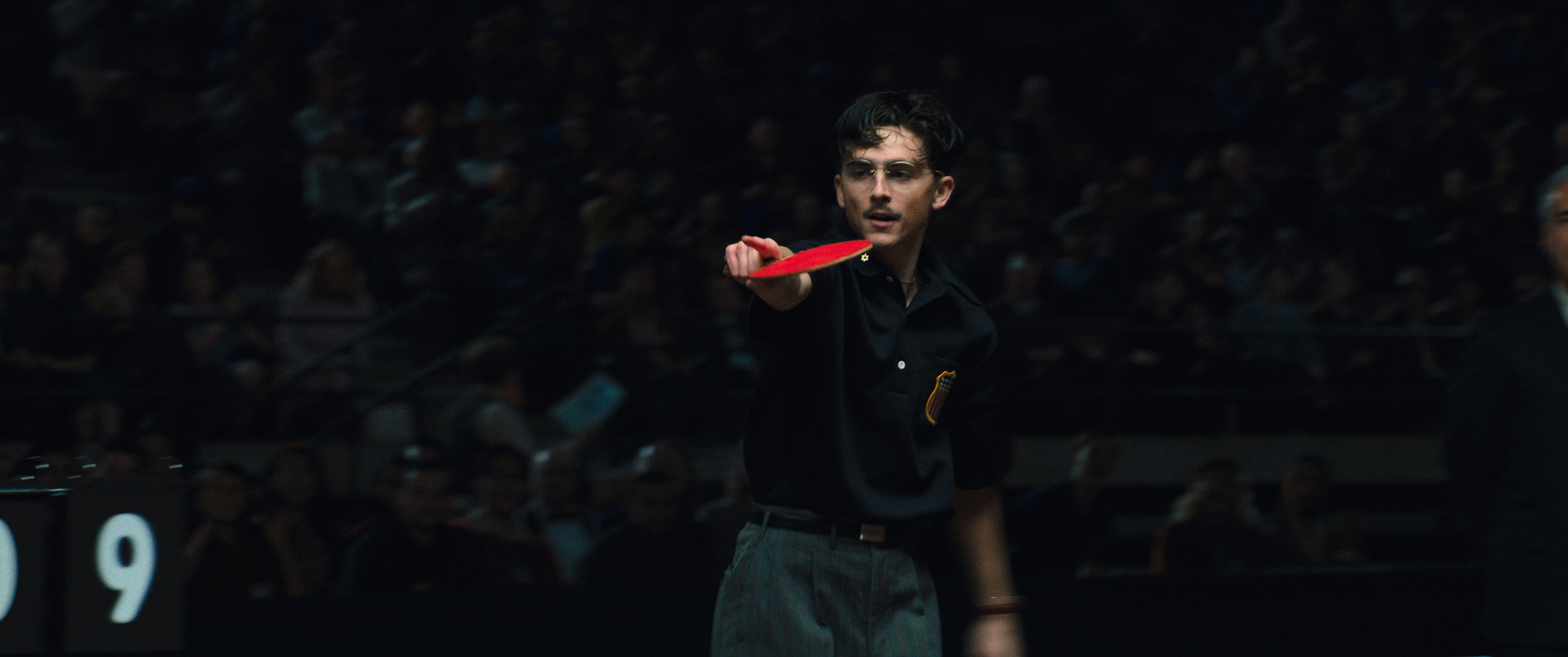 Timothee Chalamet in Marty Supreme playing table tennis in a polo shirt and glasses
