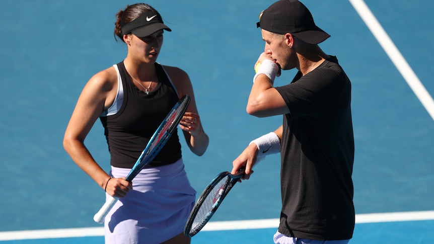 Male and female members of a mixed doubles team, wearing dark shirts, talk to each other on the court.