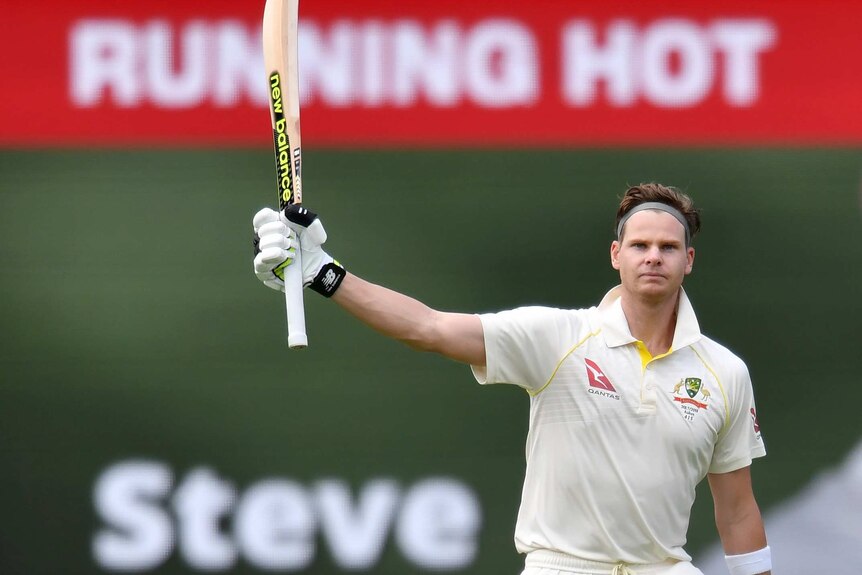 Steve Smith holds up a cricket bat with one hand with the words "Running Hot - Steve" on a sign behind him.