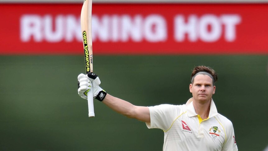 Steve Smith holds up a cricket bat with one hand with the words "Running Hot - Steve" on a sign behind him.