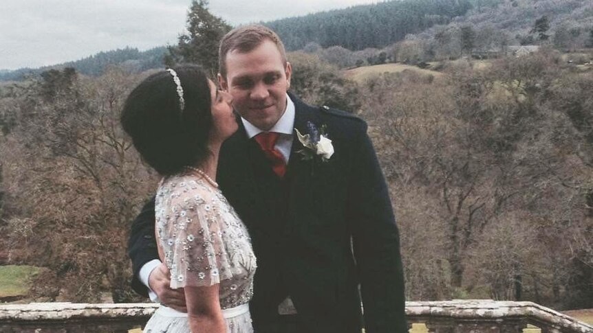 A man wearing a kilt embraces a woman wearing a white wedding dress.