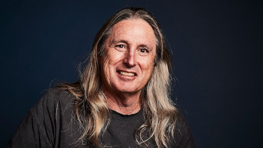 A man with long sandy-coloured hair smiles in a studio shot with a black background