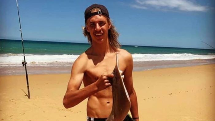 Jason Langhans is on the beach smiling and holding a stingray, standing in front of fishing poles.