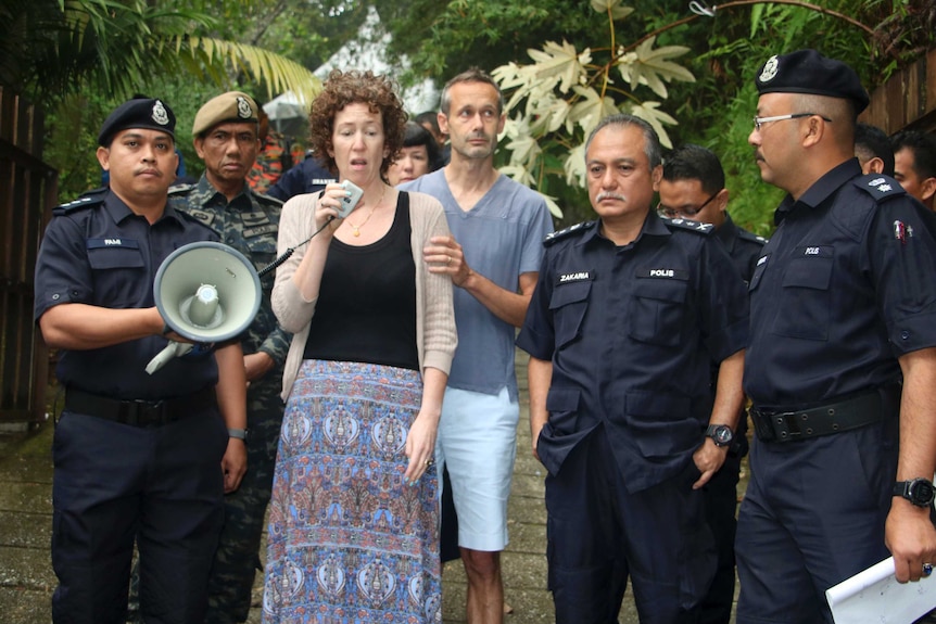 Meabh Quoirin holds a microphone in her hand as police surround her. Her husband stands behind her in the forest.