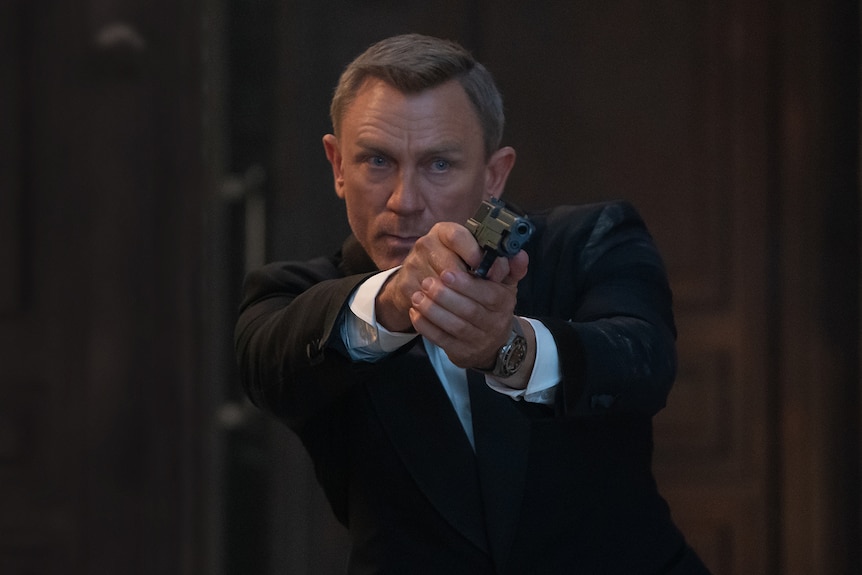 Middle-aged white man with fair cropped hair wears a black tuxedo and looks intensely while pointing a handgun