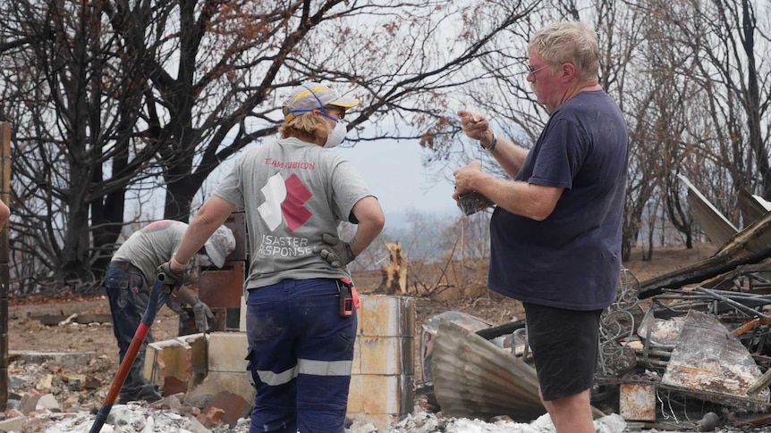 People wearing grey shirts shift through bushfire rubble, and a man looks at a small object