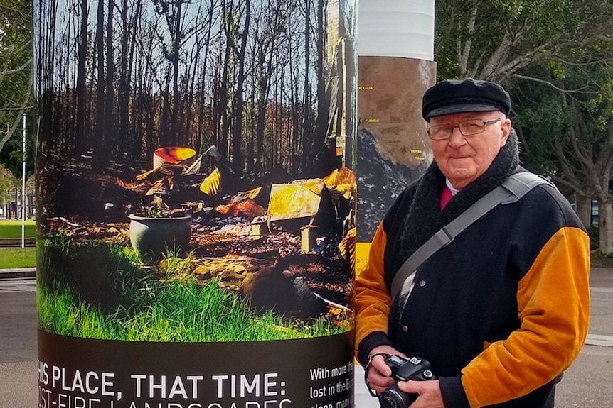 A man in his 60s stands next to a pole with a photograph of burnt bushland.