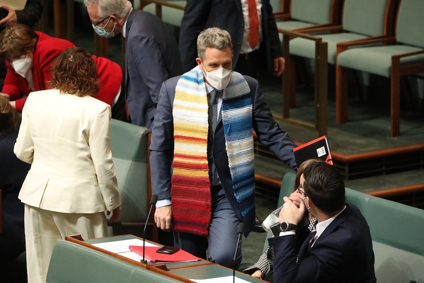 Wilson wears a long scarf with bands of red, yellow, white and shades of blue, with rows transitioning from blue to red.