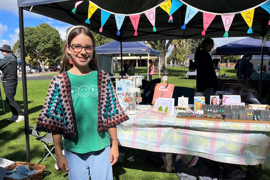 A girl in a colourful outfit stands in front of her market stall, with homemade keyrings, watercolour cards and a picture book.