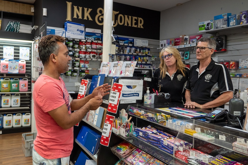 A man speaks to a man and a woman, who are standing side by side behind the counter in a newsagency.