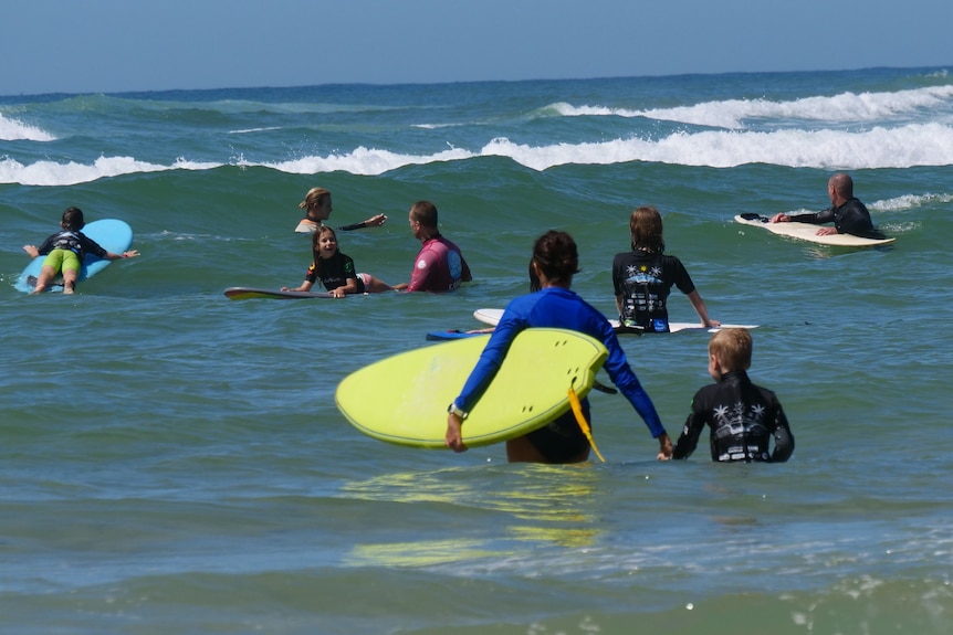 A blonde woman smiling on sunny day at the beach with four young girls around her and trees behind.