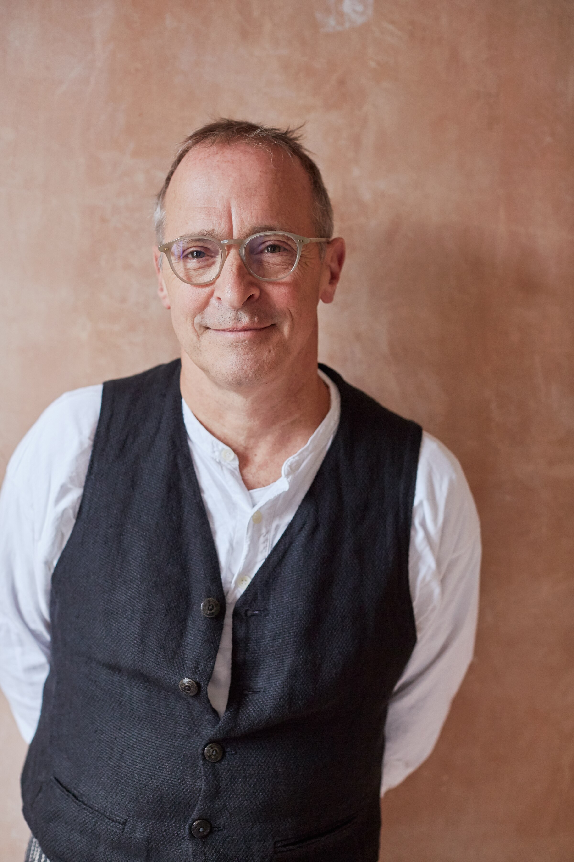 David Sedaris, a 67-year-old man with glasses, smiles slightly as he looks into the camera. He is wearing a vest.