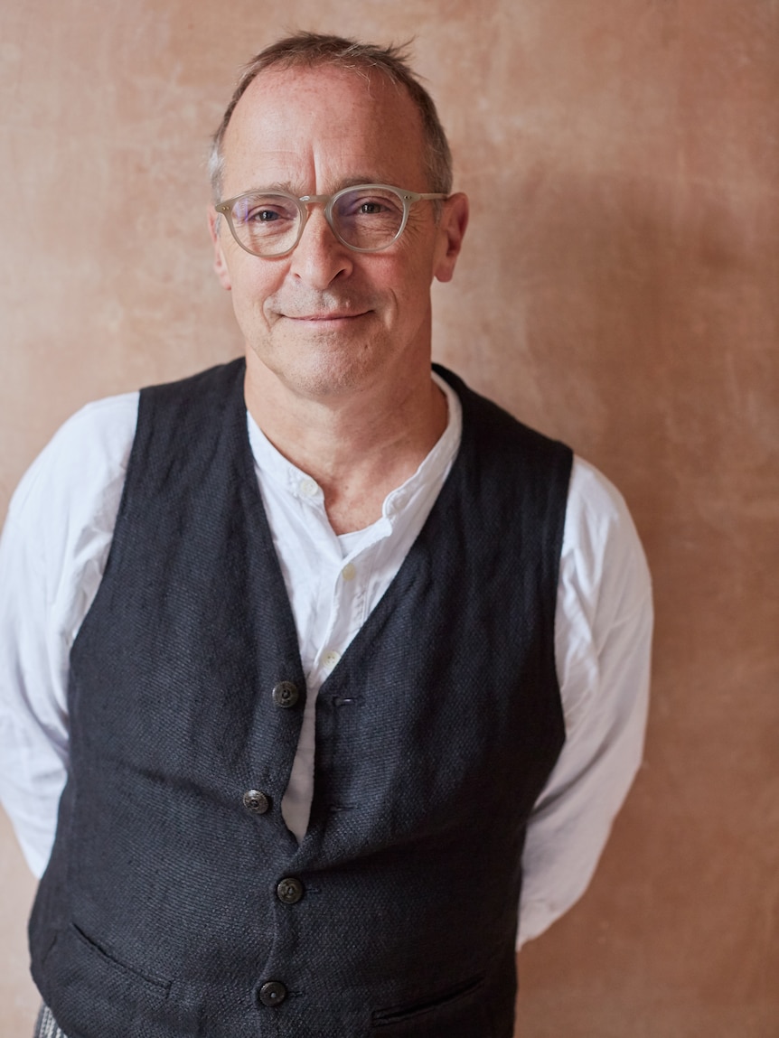 David Sedaris, a 67-year-old man with glasses, smiles slightly as he looks into the camera. He is wearing a vest.