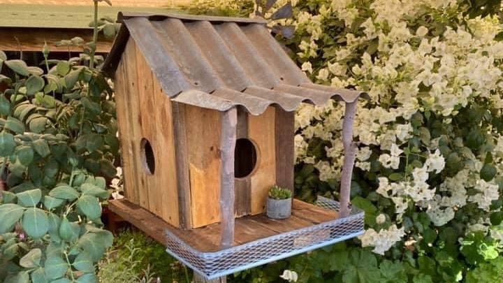 Rustic birdhouse made from old timber and tin with a cottage garden in background.