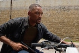 Will smith in daylight on motorbike holding gun, wall in background.