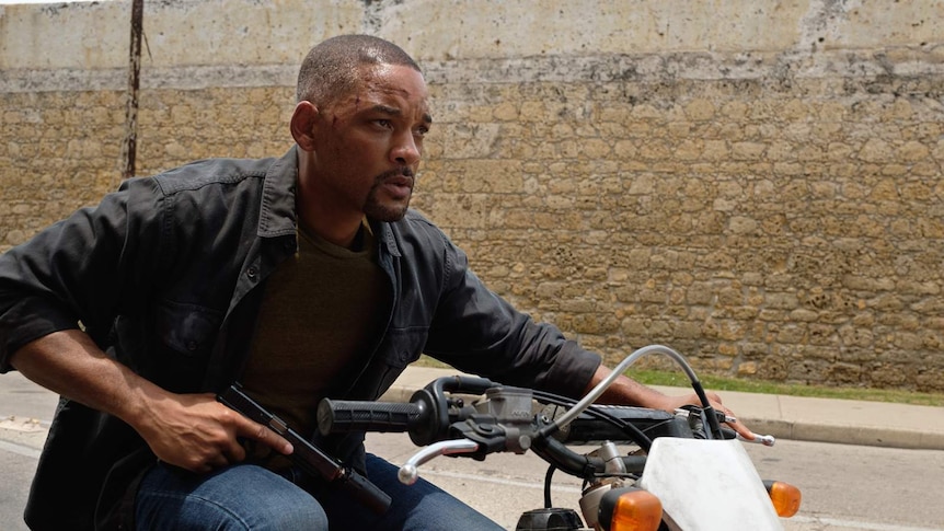 Will smith in daylight on motorbike holding gun, wall in background.