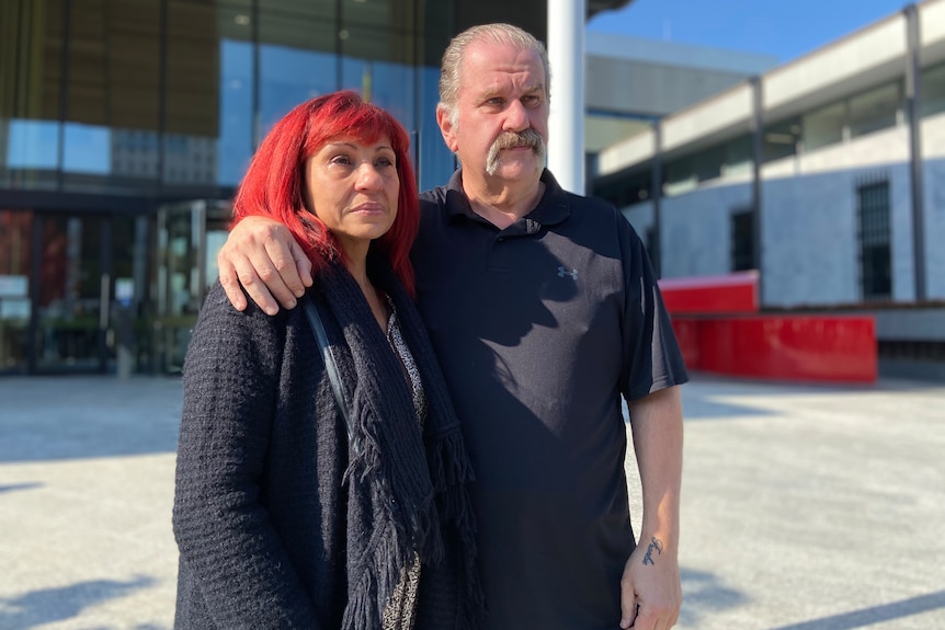 A woman with red hair and a man with a moustache stand with their arms around each other, looking into the distance.