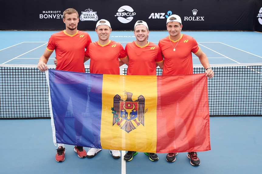 four men in Moldovan tennis uniforms pose with the Moldovan national flag on a tennis court